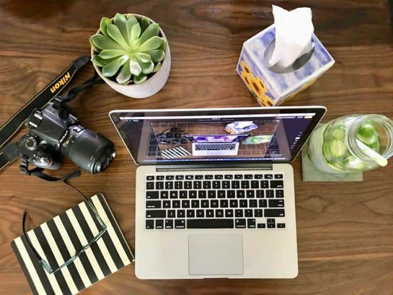 Flatlay of MacBook, succulent, camera, notebook, glasses, cucumber water and tissues.