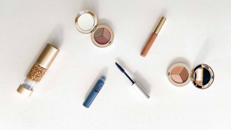 Flatlay of two eyeshadow trios, blue mascara, lipgloss, and liquid foundation.