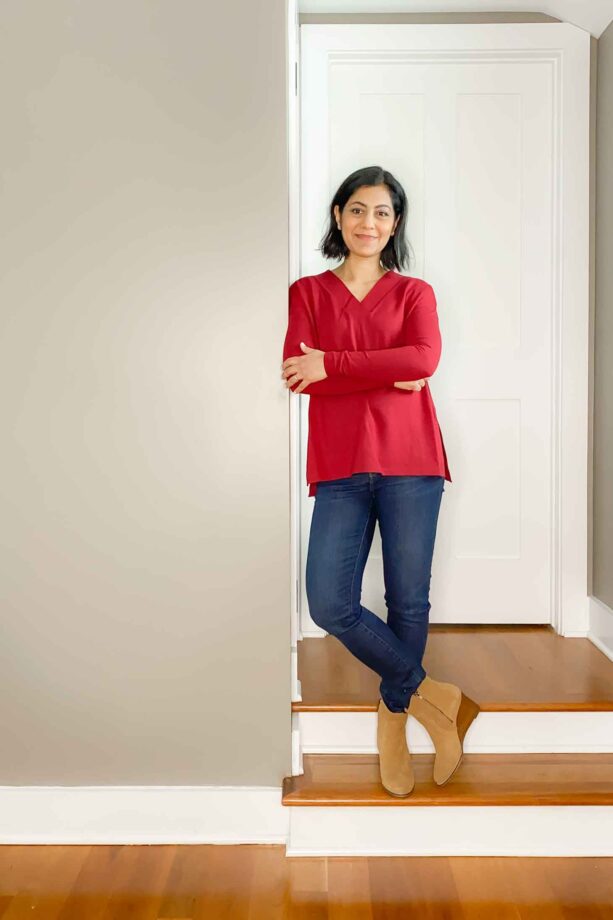 Fashionipa leaning against a wall in a red long sleeved top, blue jeans and tan boots.