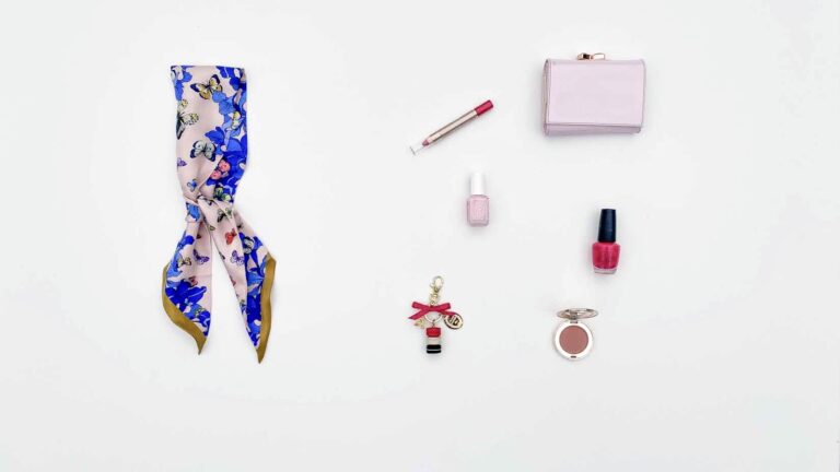Flat lay of a pink scarf with blue butterflies, lip crayon, wallet, key chain, nail polish and blush