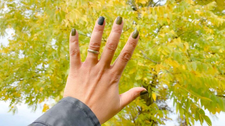 Woman's hand held up against a fall tree. She's wearing olive green nail polish on all her nails except for the ring fingernail which has denim blue polish.