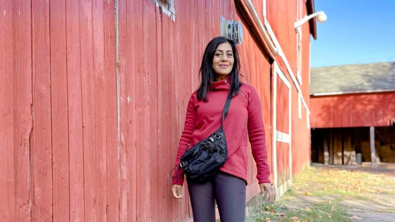 Fashionipa (Nipa) standing in front of a red barn wearing a nylon crossbody bag, a red sweatshirt, gray pants and boots.