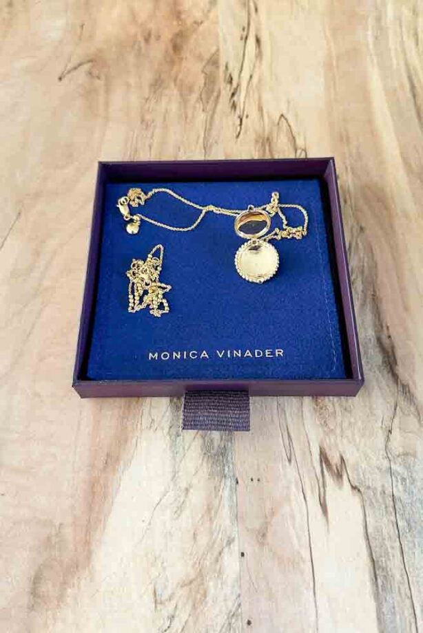 Gold chain, and round gold locket on a chain placed on a blue Monica Vinader jewelry pouch.