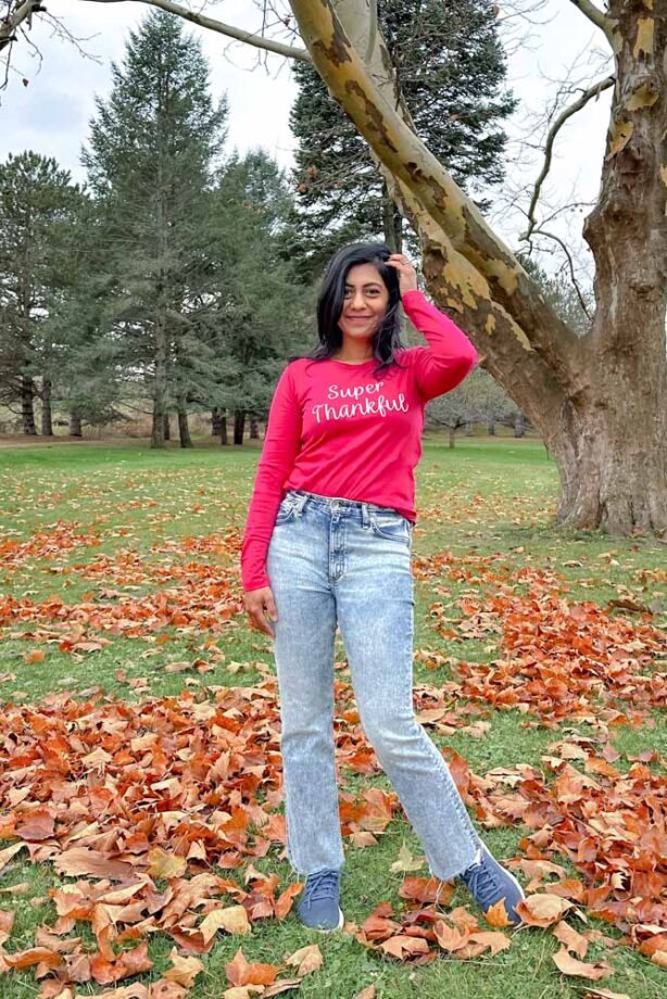 Fashionipa (Nipa) standing on fall leaves wearing a hot pink t-shirt with Super Thankful embroidered across the front, with jeans and blue sneakers