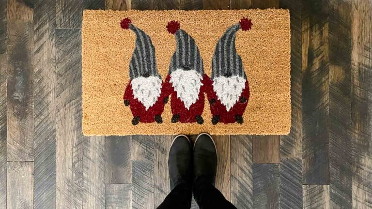 Birdseye view of a coir doormat with three gnomes in gray striped hats and red suits. Standing at the edge of the mat is a woman's black boots.