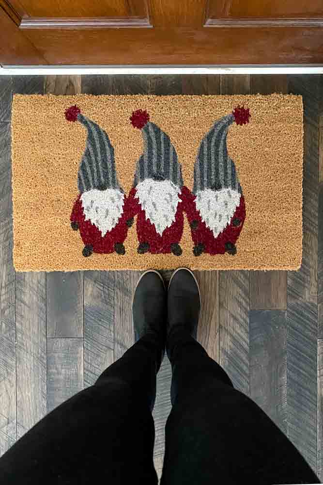 Birdseye view of a coir doormat with three gnomes in gray striped hats and red suits. Standing at the edge of the mat is a woman's black boots.