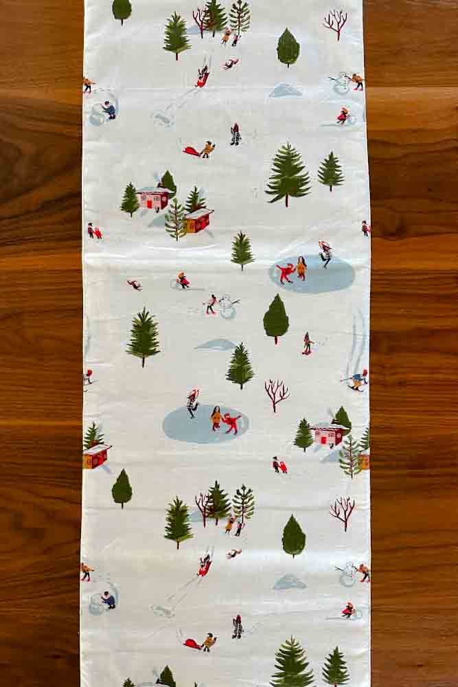 Birdseye view of a white cotton table runner with a winter scene including trees, ponds and skiers. 