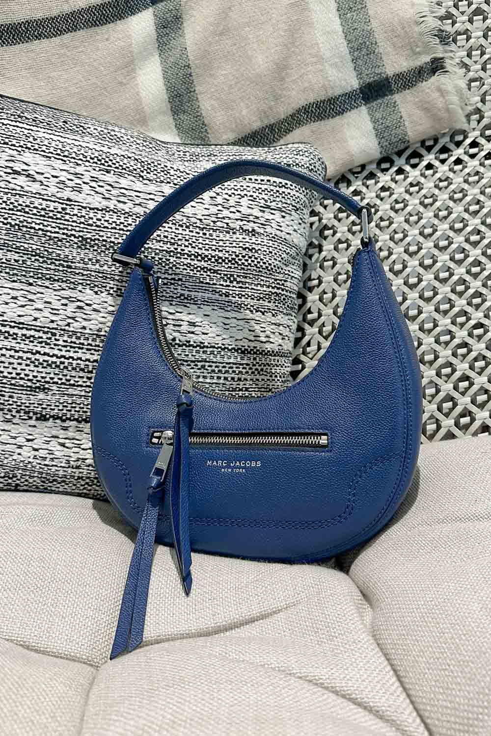 Image of a blue Marc Jacobs crescent shaped bag on a couch with black and tan printed cushions.