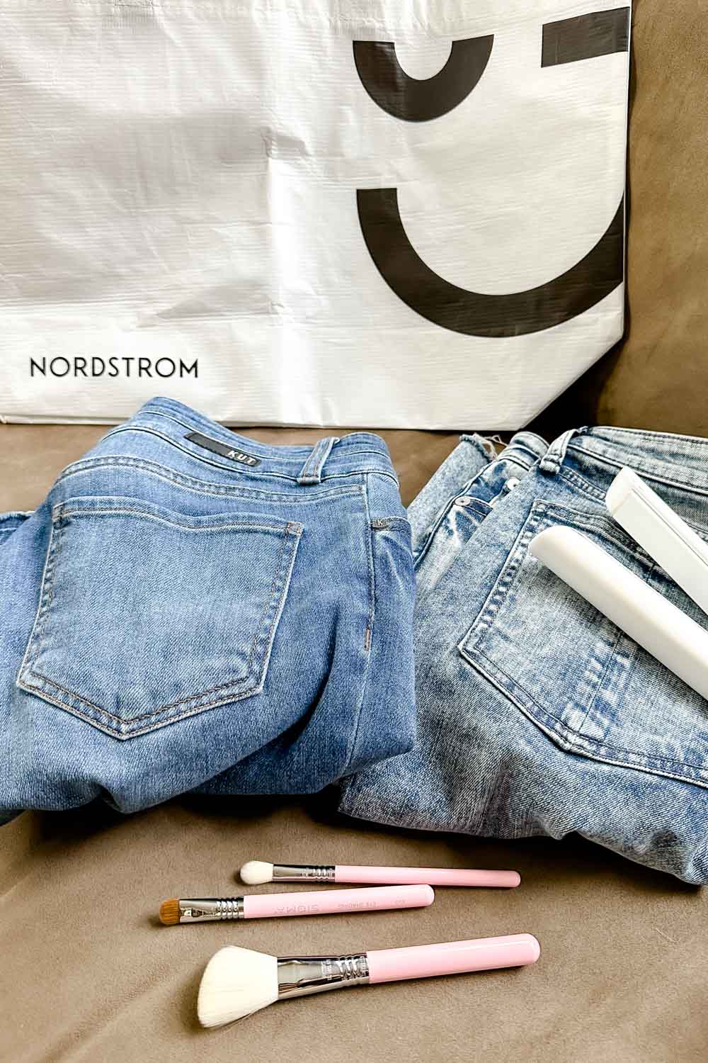 A Nordstrom shopping bag, two pairs of jeans, a flat iron and three makeup brushes on a tan couch