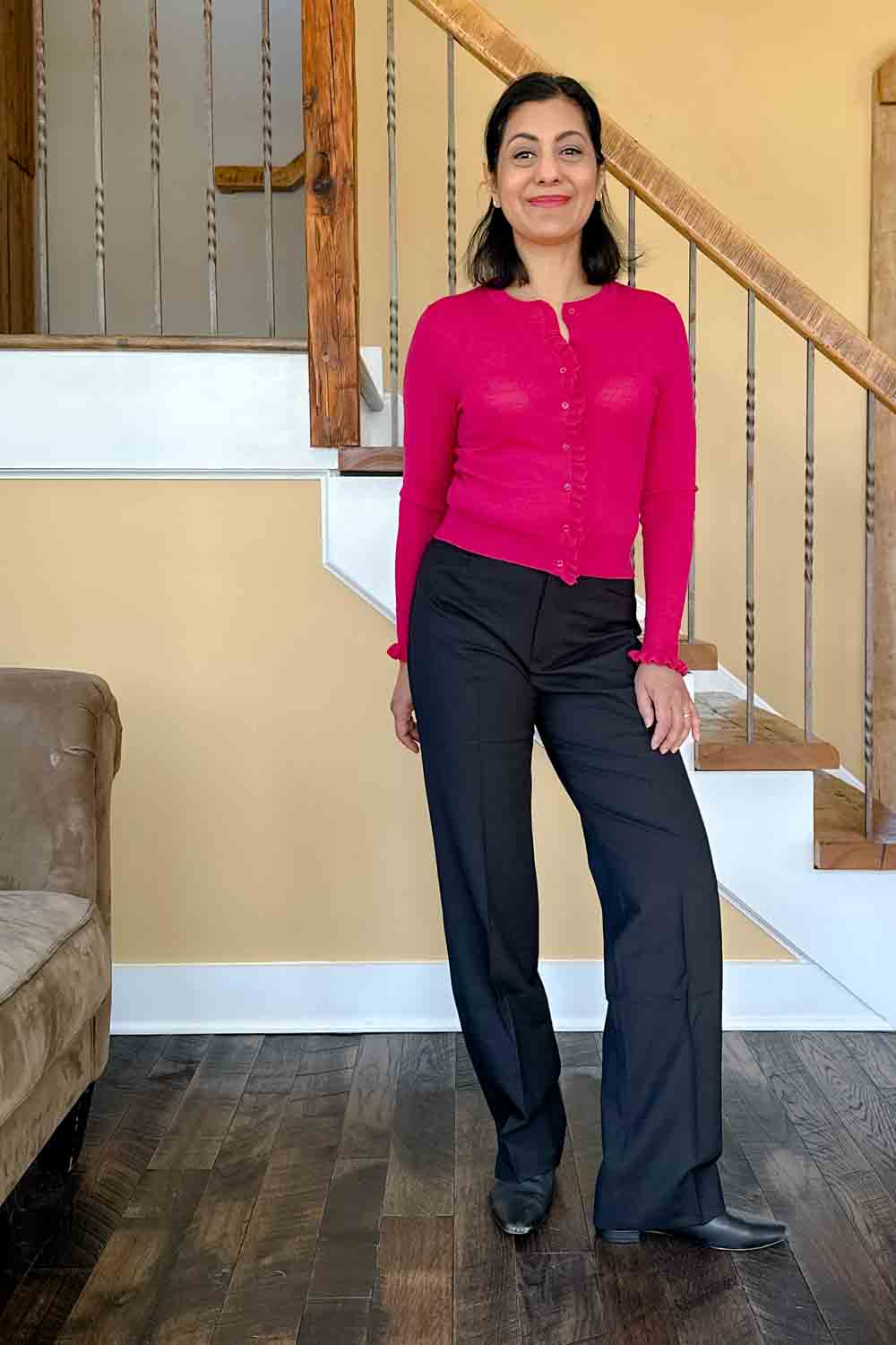 Fashionipa (Nipa) standing in front of a staircase wearing a pink sweater and straight leg black pants.