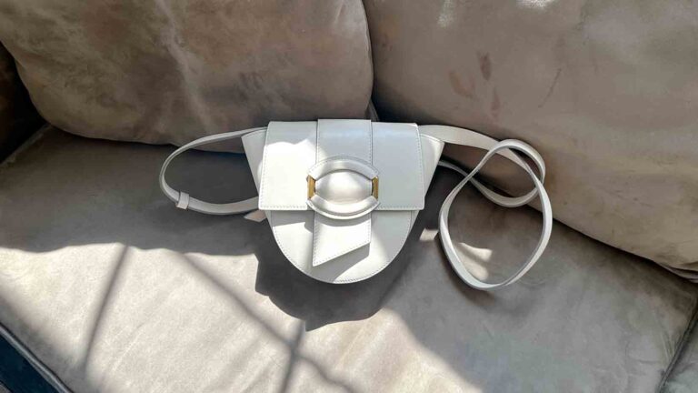 Image of a stone crossbody bag with an oval buckle on a tan couch.