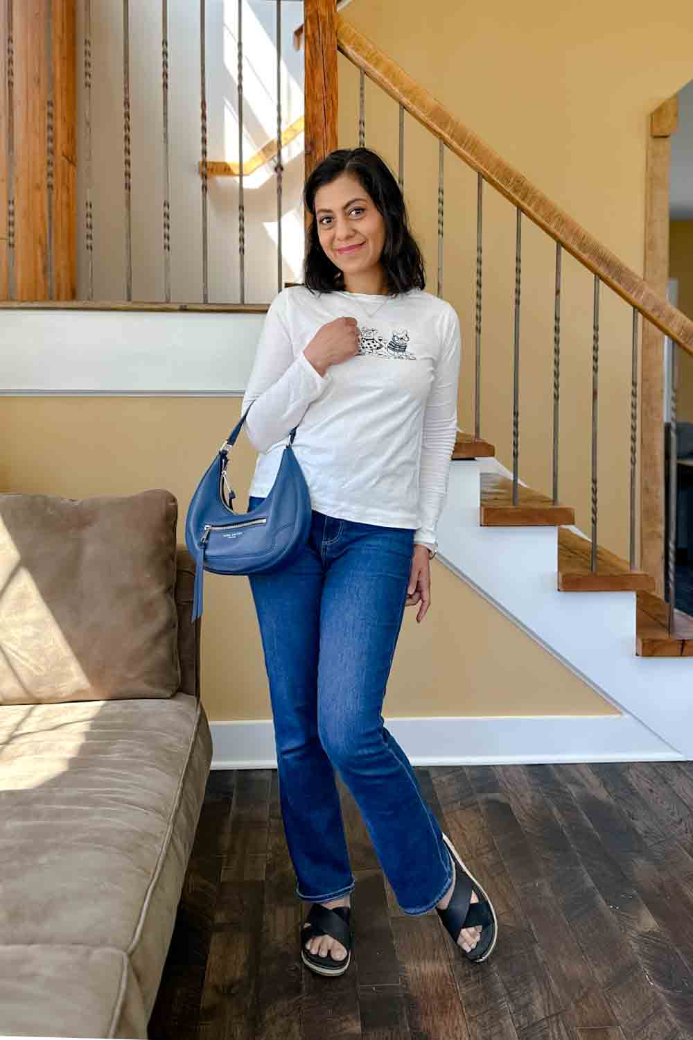 Fashionipa (Nipa) standing in front of a staircase in a white t-shirt, blue jeans, black sandals, carrying a blue crescent bag...