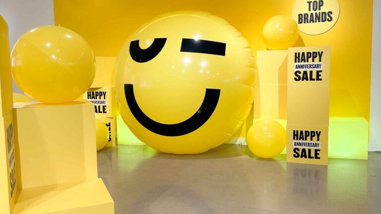 Photo of a large inflatable ball in yellow with a winking face next to Anniversary Sale signs (at Nordstrom)