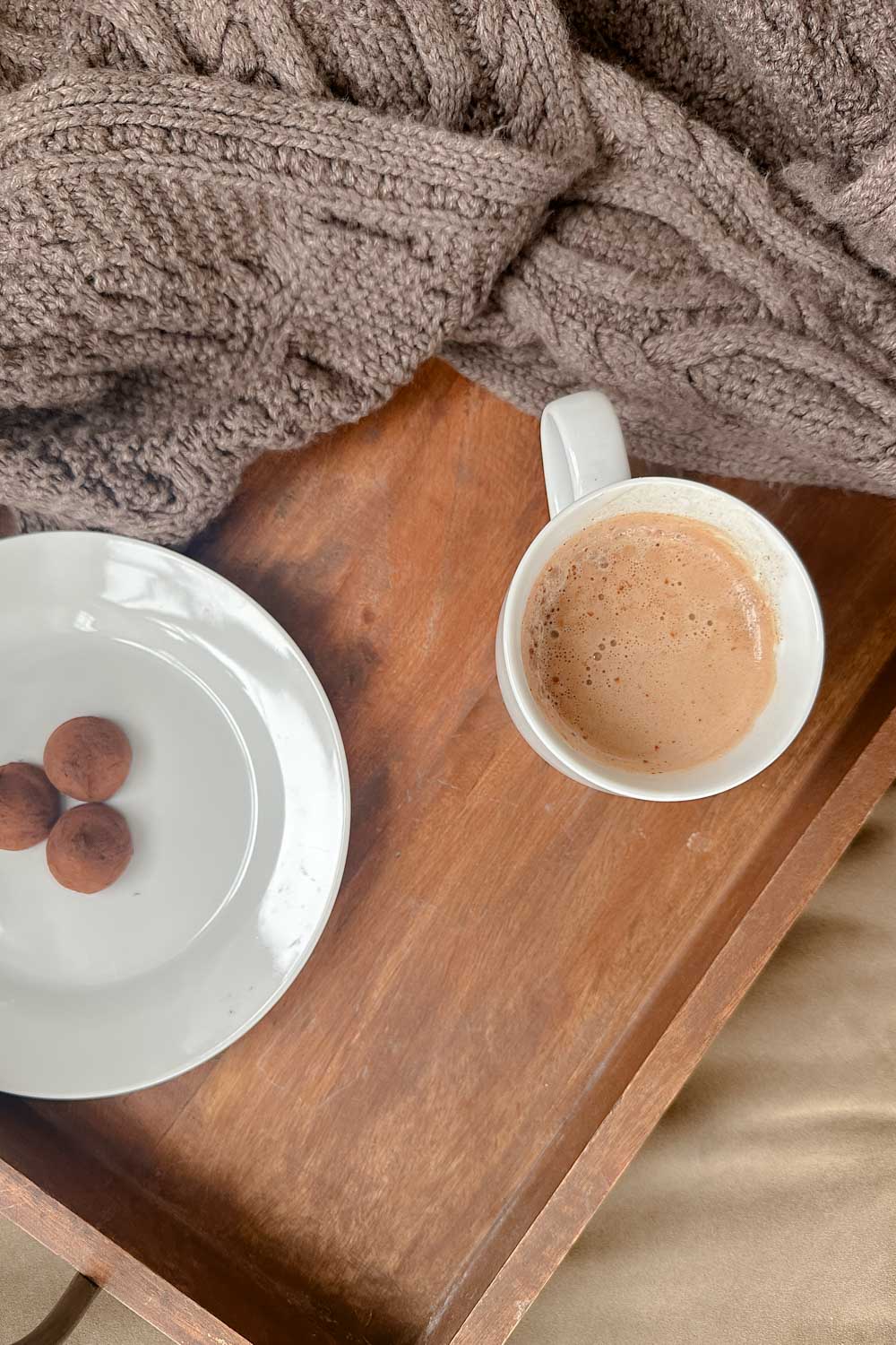 A cup of mocha in a white mug, a plate of chocolate truffles and a knit throw on a wooden tray.