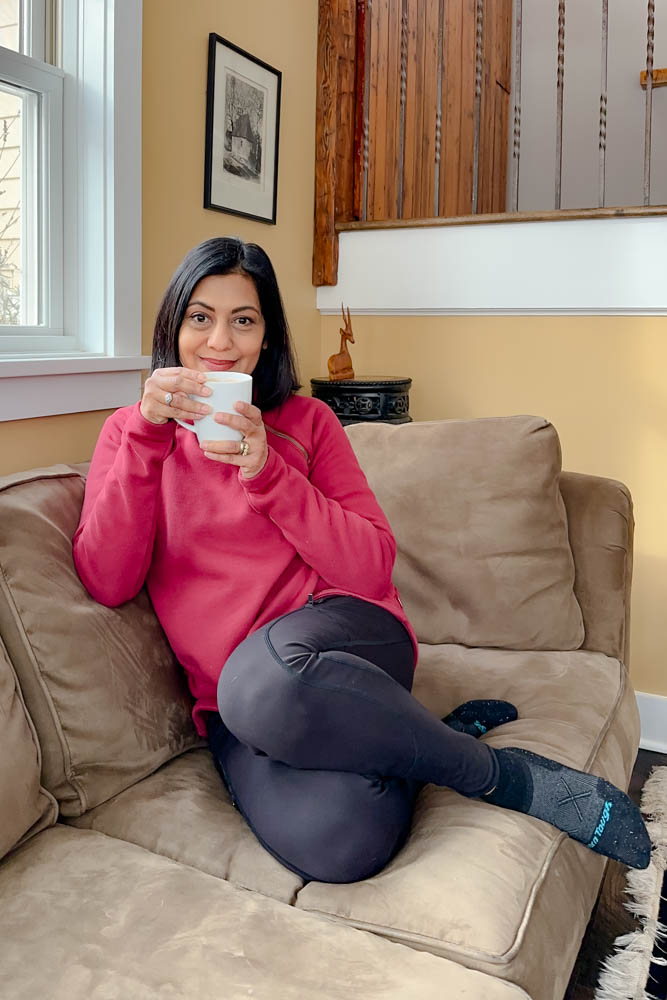 Fashionipa (Nipa) sitting on a couch wearing a red sweatshirt, black leggings and black socks, holding a white mug.