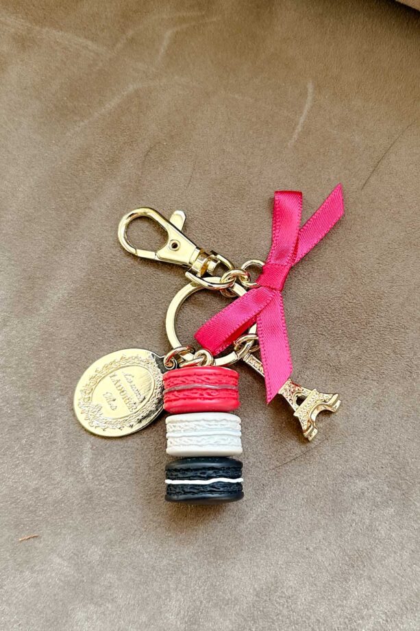 Cream Coach handbag Charm that features stack of macarons, a pink ribbon, and a miniture Eiffel tower.