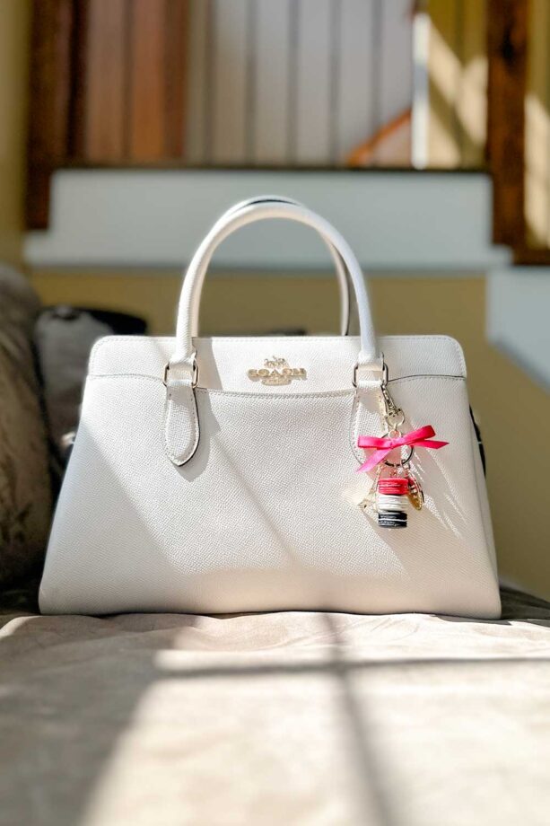 Cream Coach handbag with a charm that features stack of macarons, a pink ribbon, and a miniture Eiffel tower.