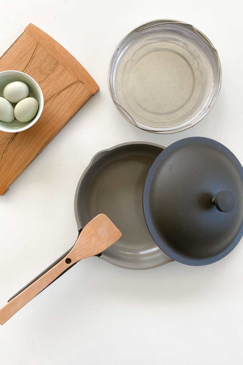 Birds-eye-view of the Always Pan with wooden spatula and lid, steamer basket, a cutting board and a bowl of eggs.