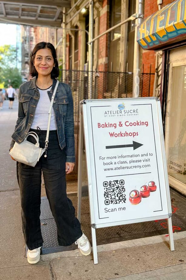 Fashionipa (Nipa) standing in front of the Atelier Sucré sandwich board. the board reads Baking & Cooking Workshops. For more information, and to book a class, please visit www.ateliersucreny.com