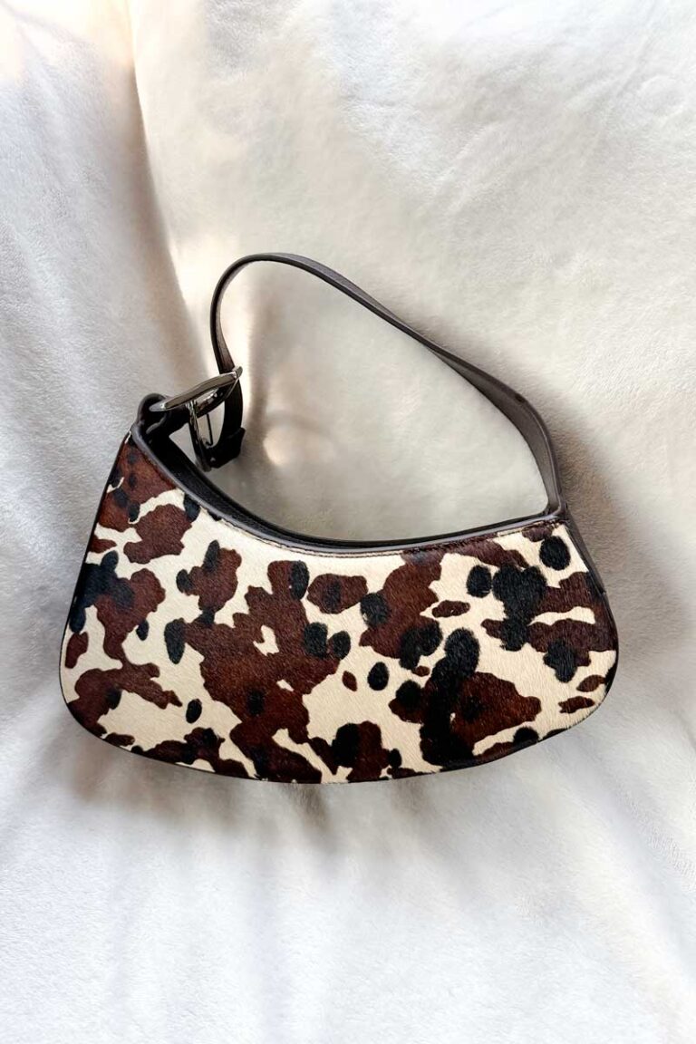 An asymmetrical handbag in a brown, black and white cow print (on a white blanket)