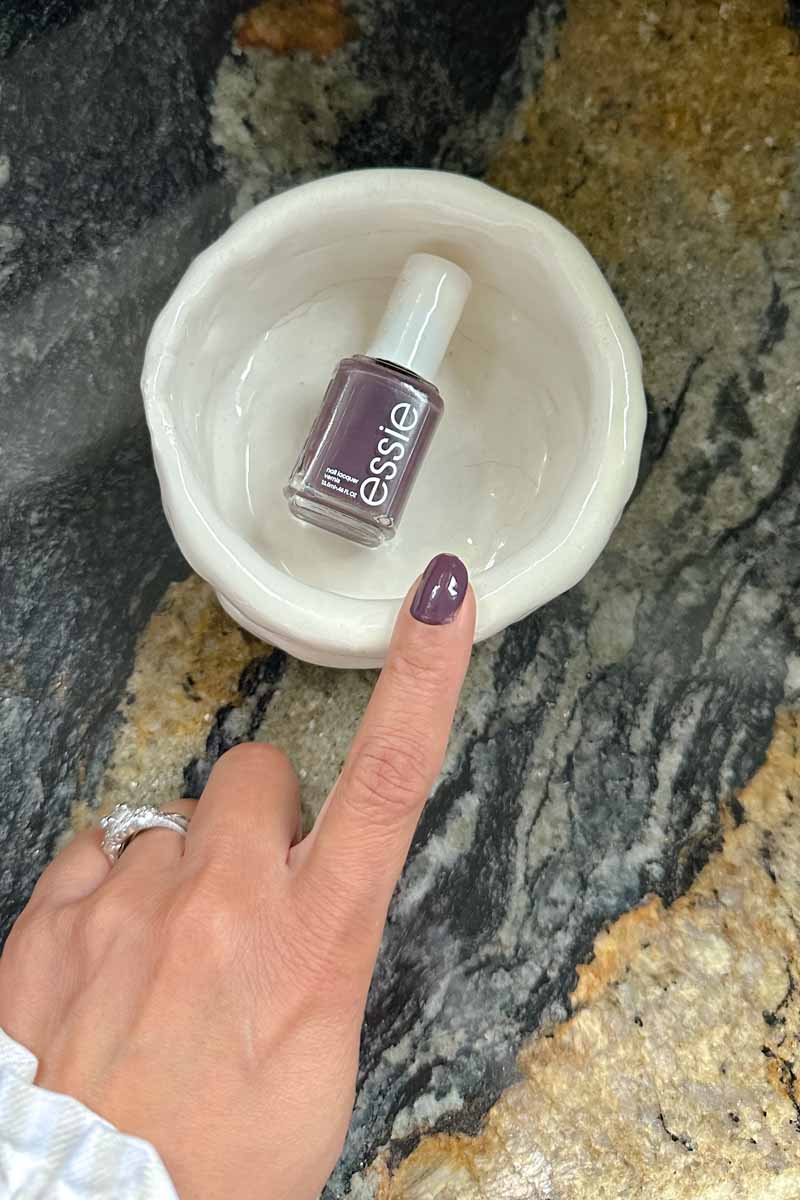 A woman's hand with the index finger pointing to a nail polish bottle in a white bowl - the color of the polish is a deep purple.