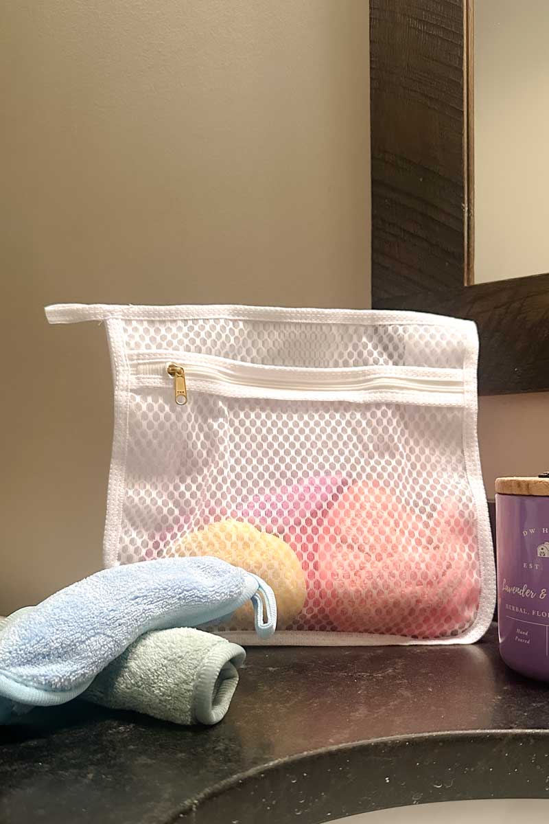 Mesh laundry bag with microfiber makeup removing cloths inside. Two more cloths are in front of the bag, on a bathroom countertop.  