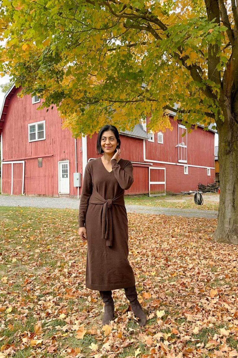 Fashionipa (Nipa) wearing a brown sweater dress and boots standing in front of a red barn with fall leaves on the ground.