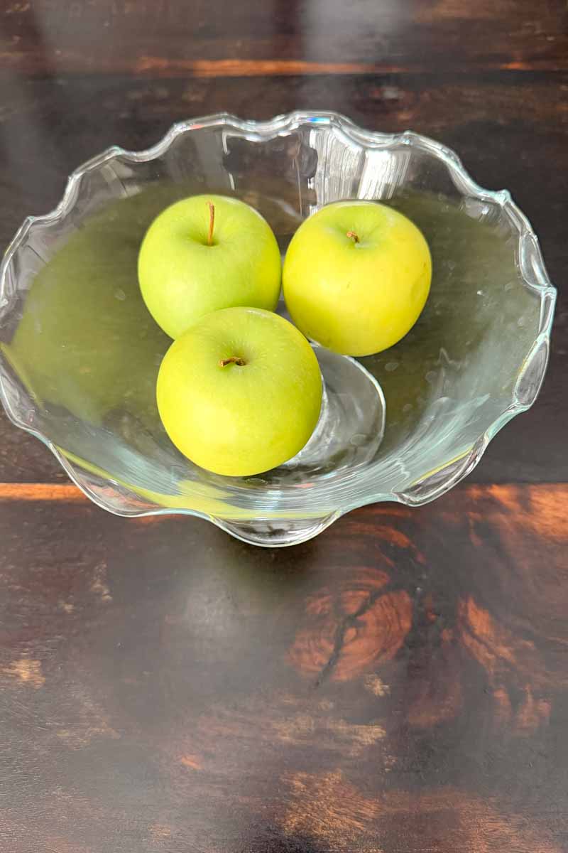 Footed glass bowl with ruffled edges containing three green apples.