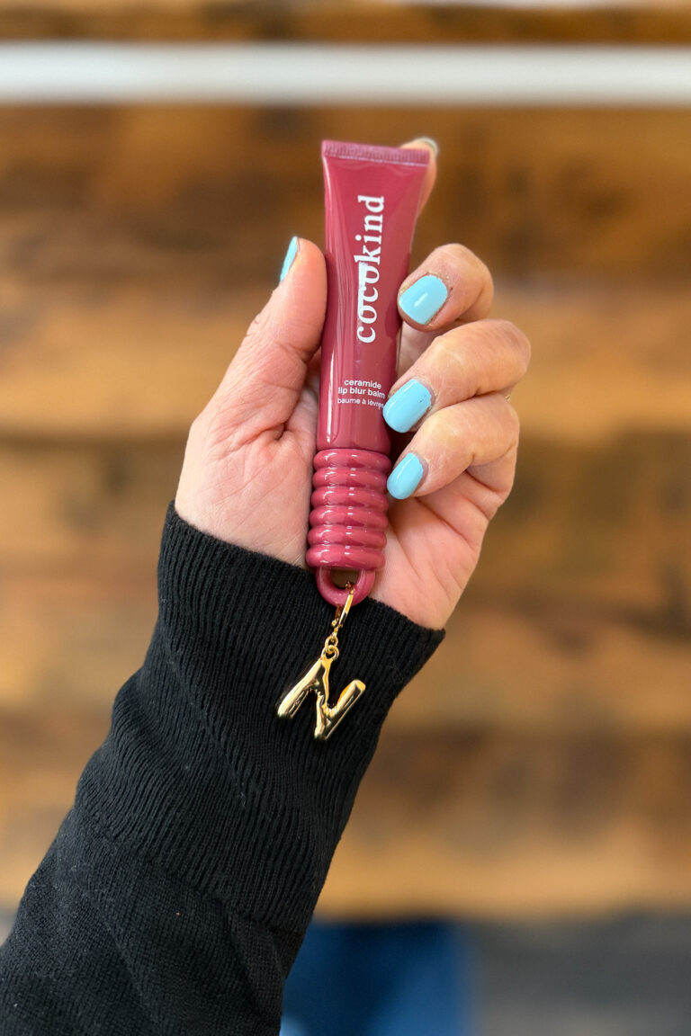 Woman's hand holding Cocokind's lip balm in Be Bold with a gold initial charm (letter N) attached.