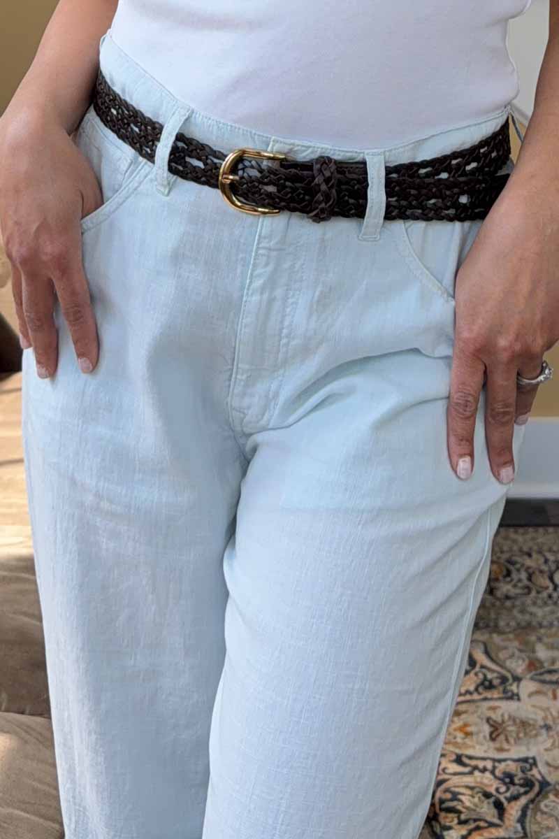 A woman wearing blue pants, a white t-shirt and a dark brown open weave leather belt. 