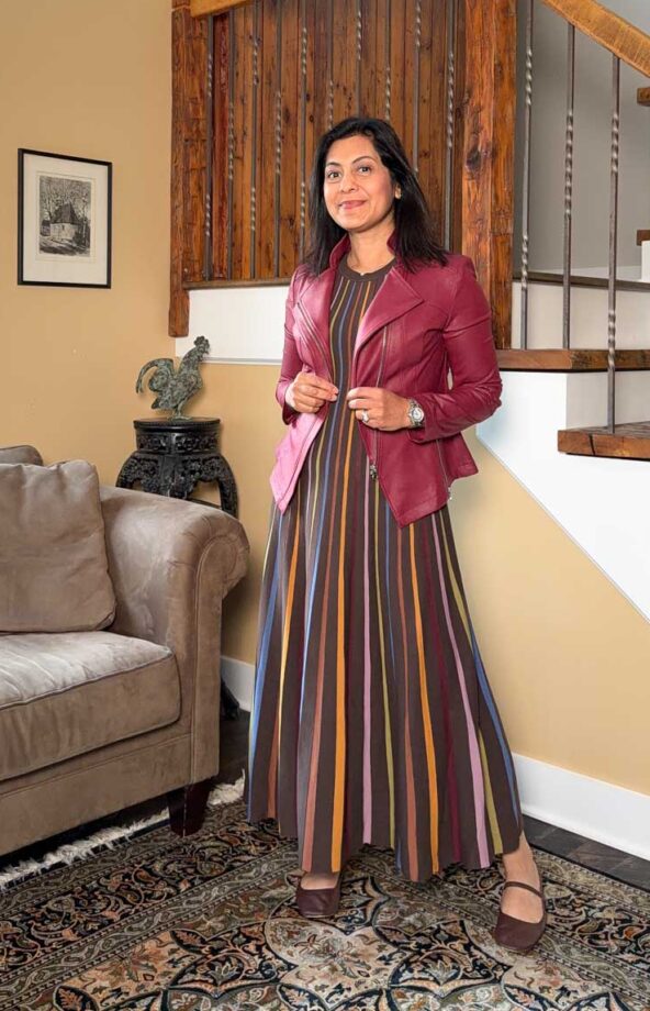 Fashionipa (Nipa) wearing a striped knit maxi dress and burgundy moto jacket, stadning next to a couch and staircase.